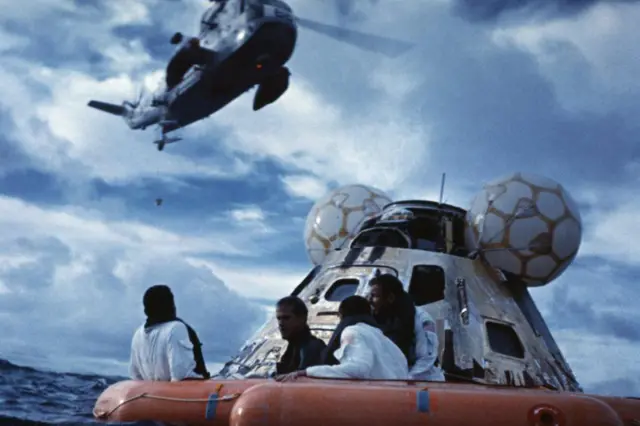 Astronautas del Apolo 13 esperan ser rescatados en una balsa. La cápsula espacial se ve detrás de ellos y un helicóptero sobrevuela.