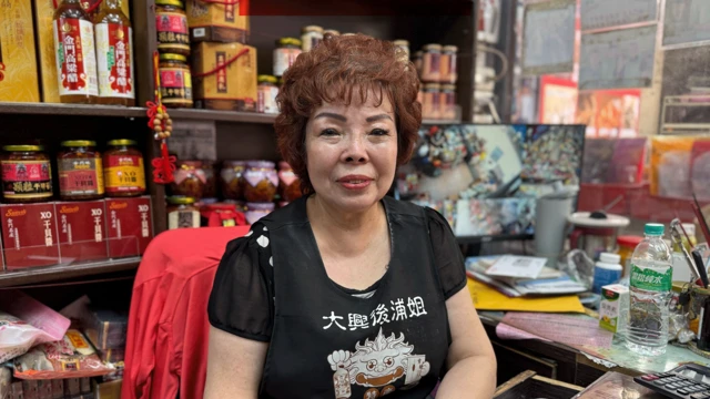 Yang Peiling, una mujer con pelo corto y castaño, sentada en una tienda de alimentos, con una camiseta de manga corta negra que tiene un diseño y símbolos orientales