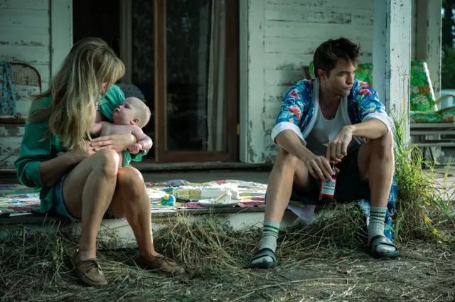 En una escena de la película Die My Love, Jennifer Lawrence carga a un bebé mientras está sentada en una acera. A su izquierda, indiferente, Robert Pattinson sostiene entre sus manos una cerveza.