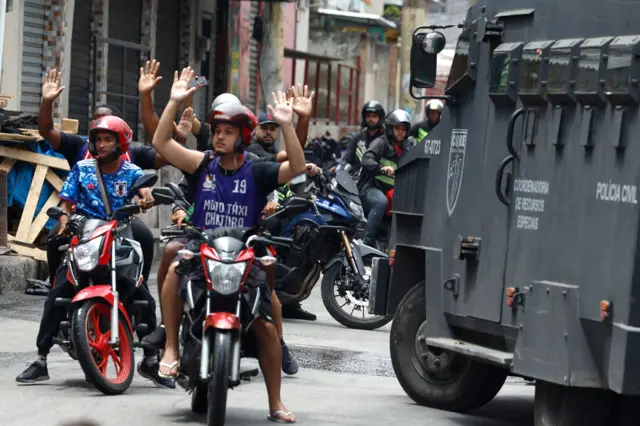Motociclistas con las manos levantadas al lado de un camión de la policía.