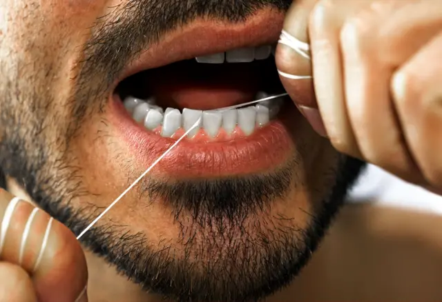Imagen cerrada de la boca y cara de un hombre con barba que coloca hilo dental entre los incisivos