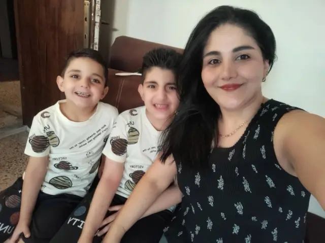 Randa Dowiar en primer plano, con un vestido de tirantas, se hace un selfie junto a sus dos hijos, vestidos igual. 