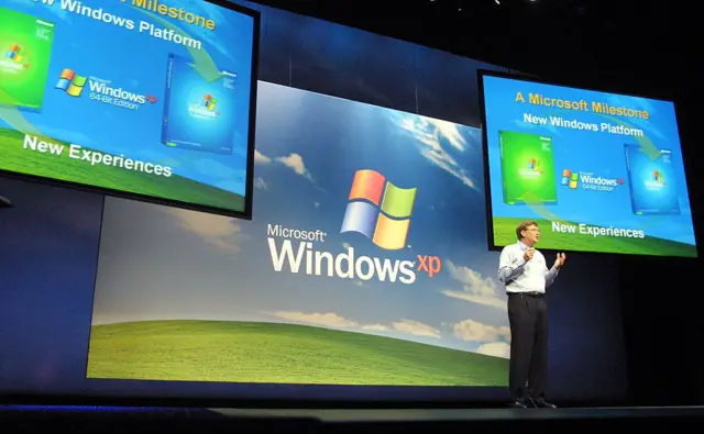 Bill Gates, vistiendo camisa blanca y pantalón de traje negro, presenta el sistema operativo Windows XP frente a unas pantallas que muestran el logo de Microsoft y el icónico fondo de pantalla con la colina verde y el cielo azul.