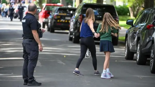 Un policía en medio de la calle supervisa mientras dos chicas avanzan de la mano. Todos aparecen de espaldas.