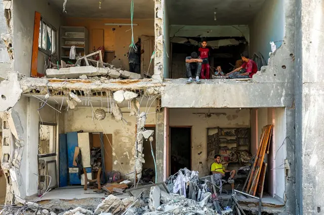 Un hombre y unos niños están sentados en los cuartos expuestos en un edificio serveramente destruido. La fachada entera del edificio se ha desplomado, dejando el interior de cuatro cuartos visible, con escombros esparcidos por todas partes   