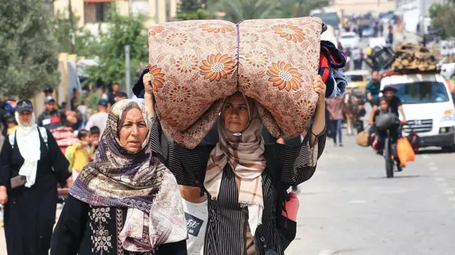 Dos mujeres con pañuelos en la cabeza caminan por una carretera, huyendo de la zona de Rafah el 28 de mayo de 2024. Una de ellas lleva un gran colchón enrollado sobre la cabeza. Detrás de ellas hay más personas caminando con sus pertenencias, vehículos repletos de pertenencias y un hombre y un niño en una bicicleta cargada.