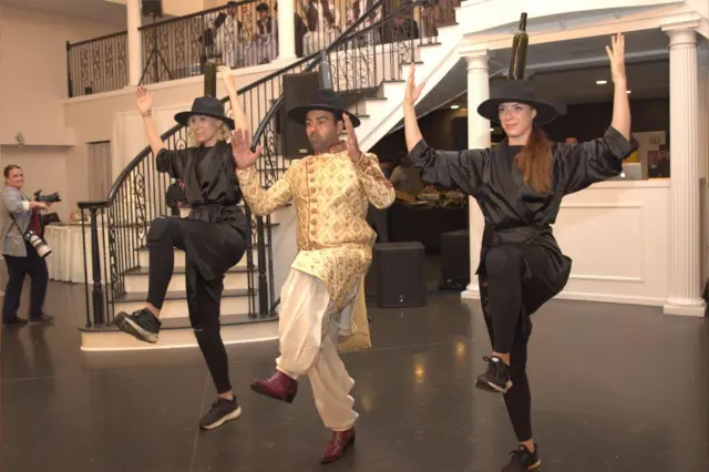 Safi y Sammi bailan junto a otra mujer al pie de una escalera interior. Las tres llevan sombreros de ala ancha con botellas sujetas a la parte superior. Tienen la pierna izquierda en el aire y las manos separadas. La gente se agolpa en el suelo y observa desde un balcón.