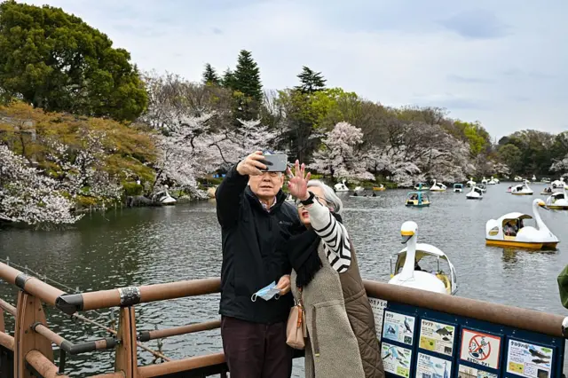 Una pareja de ancianos se toma un selfie con su móvil frente a un estanque lleno de barcos con flores de cerezo en el parque Inokashira en Tokio, Japón.