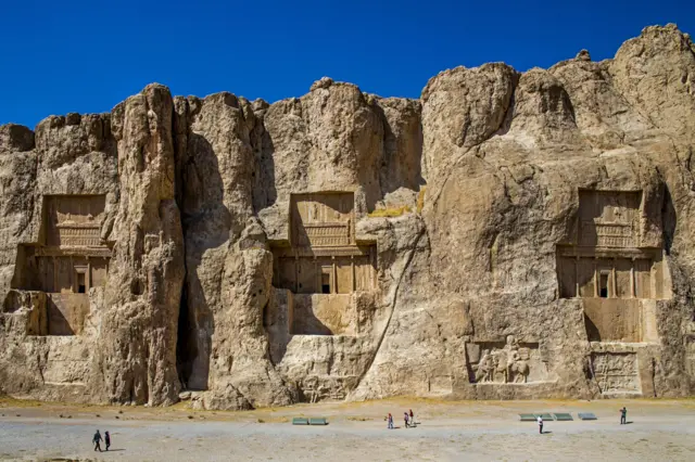 Una antigua necrópolis persa está excavada directamente en una enorme pared rocosa. La piedra caliza presenta portales de formas intrincadas. Los turistas pasan caminando bajo un cielo azul intenso.