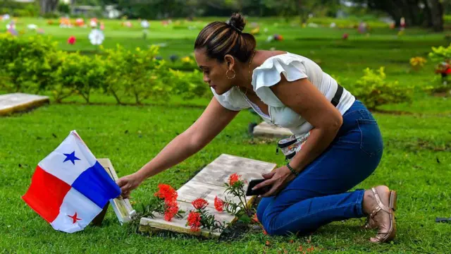 Una joven con una blusa azul, pantalones vaqueros y sandalias coloca una bandera panameña en una tumba en un cementerio con un césped bien cuidado. 