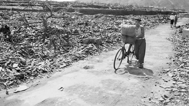 Un civil japonés empuja su bicicleta cargada por un sendero que fue despejado de escombros tras el ataque del 9 de agosto de 1945. A ambos lados del camino, escombros, metal retorcido y tocones de árboles nudosos cubren el área.