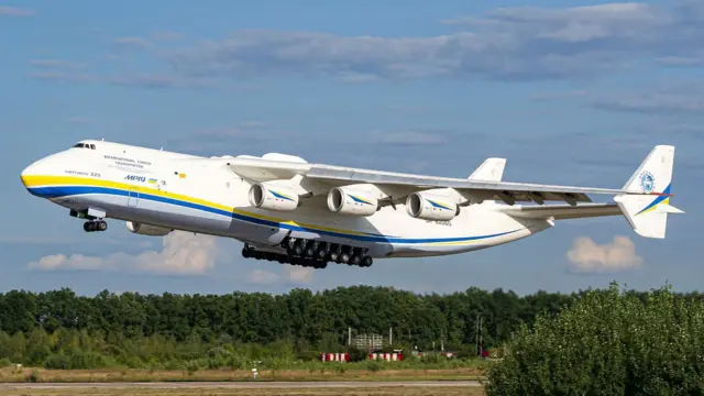 Un gran avión blanco adornado con rayas amarilla y azul a lo largo del fuselaje despega de una pista bordeada de árboles, bajo un cielo azul con unas nubes blancas. El avión tiene seis turbinas y un enorme tren de aterrizaje de 14 ruedas en el medio