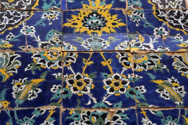 Los azulejos islámicos, con intrincados patrones, se agrietan bajo tensión. Presentan motivos florales en azul, amarillo, negro y blanco.