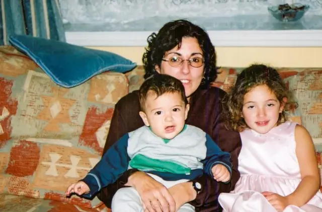 Sarah sostiene a Eric en su regazo mientras Aviva está sentada a su lado. Ambos son niños.