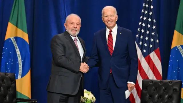 Lula y Joe Biden se dan un apretón de mano mientras miran a la cámara. Ambos visten trajes oscuros. En el extremo izquierdo se ve la bandera de Brasil y en el derecho, la de Estados Unidos.