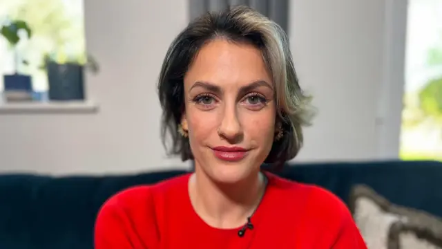 Madelaine Thomas está sentada en un sofá azul marino, sonriendo a la cámara. Lleva un vestido rojo de manga corta. Tiene el pelo corto y oscuro con una mecha rubia al frente.