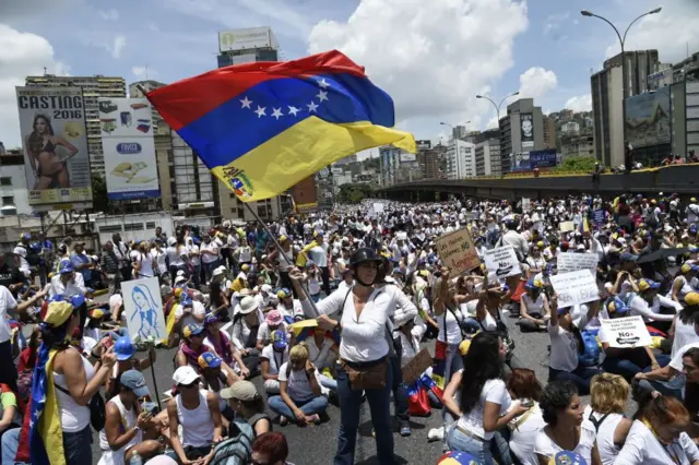 Un montñon de mujeres, de pie o sentadas, con camisetas de color blanco, ataviadas con banderas venezolanas, cascos de motorizados, y llevando carteles de protesta, en medio de una de las principales vías de Caracas. 