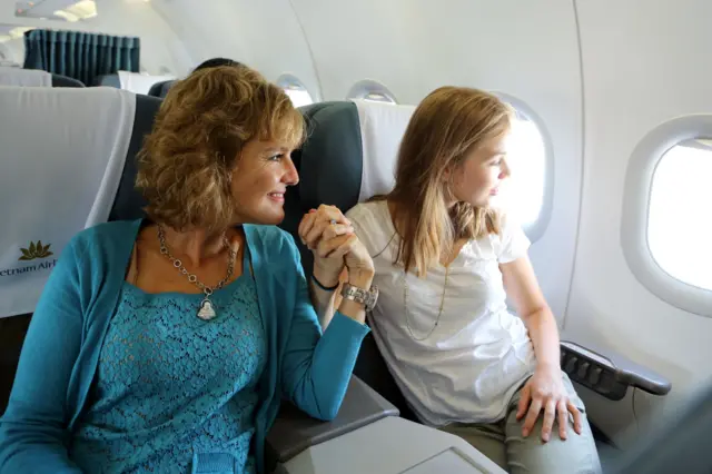 Annette y su hija agarradas de la mano sentadas en un avión mirando por la ventana.