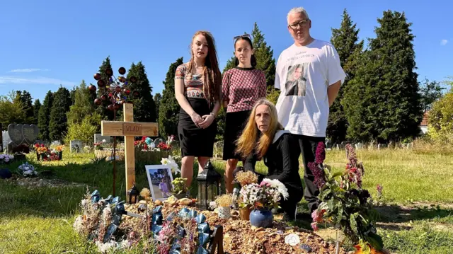 La familia de Vlad, Mia, Masha, Anna y Graham, junto a su tumba en Southampton, que está cubierta de tributos y marcada por una cruz de madera hecha a mano con su nombre