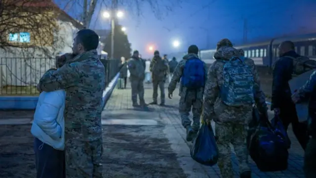 Un hombre con uniforme militar abraza a una mujer en una estación de tren. A su lado, pasan otros militares cargados con mochilas y otros bultos.
