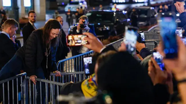 María Corina Machado saltando unas barricadas para acercarse a un grupo de personas que le esperan con celulares en mano. Lleva un abrigo negro, un pantalón de mezclilla y varios rosarios le cuelgan del cuello. 