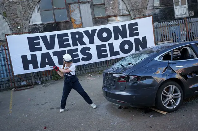 Una mujer golpeando un Tesla con un martillo, con un gran letrero atrás que dice 