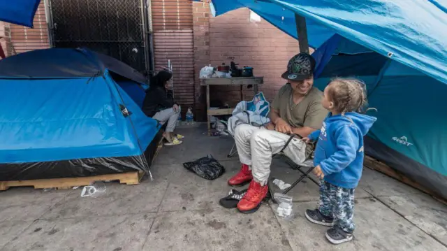 Jhon Valencia juega con Thian, de 2 años, con su madre Katherine González está sentada en una tienda de campaña al fondo, en Towne Avenue en Skid Row, en Los Ángeles, el jueves 25 de abril de 2024. (Myung J. Chun / Los Angeles Times a través de Getty Images)