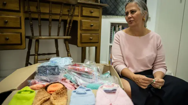 La foto muestra una caja de cartón llena de pequeñas prendas tejidas y a su lado está sentada la neonatóloga Lilia Embiruçu.