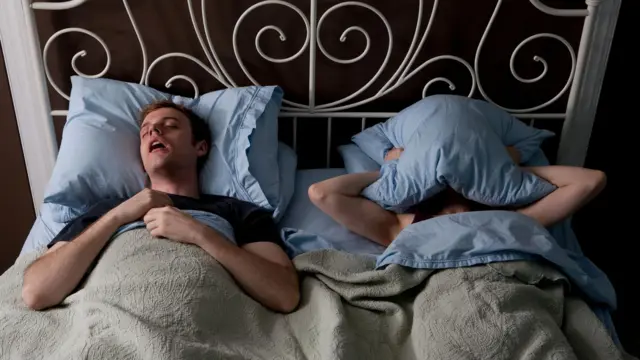Una pareja en una cama. El hombre ronca con la boca abierta y la mujer se tapa la cara con una almohada para no escuchar