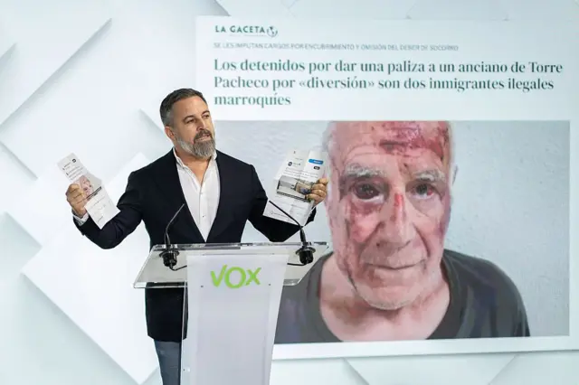 El líder de Vox, Santiago Abascal, en una rueda de prensa con la imagen del anciano agredido de fondo.