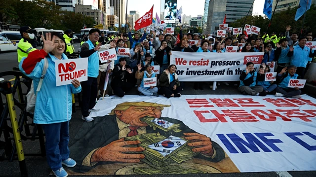 Varias personas con pancartas anti-Trump se congregan alrededor de una pancarta que muestra una caricatura de Trump vomitando dinero.