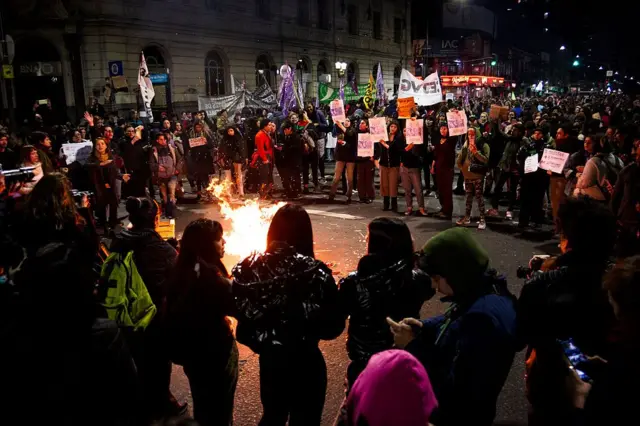 Varias personas en una manifestación hacen un corro y en el centro hay un fuego. Algunas llevan pancartas. 