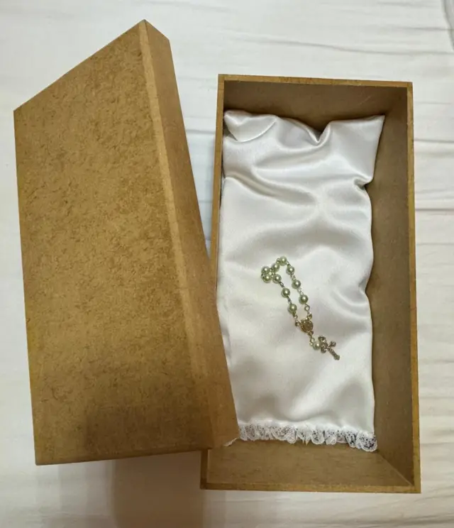 Una caja de madera más pequeña que una caja de zapatos, abierta, que contiene un pequeño rosario sobre un cojín blanco.