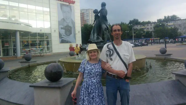 Serguéi y Tatiana en una plaza pública de Moscú, con una fuente y una estatua detrás. Ella lleva un vestido azul y blanco, y él, una camiseta blanca y vaqueros.