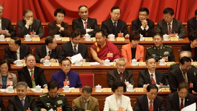El Panchen Lama, designado por China y vestido con el atuendo rojo tradicional de los monjes tibetanos, se sienta entre hombres y mujeres con trajes y uniformes militares en filas de asientos en una sala de conferencias.
