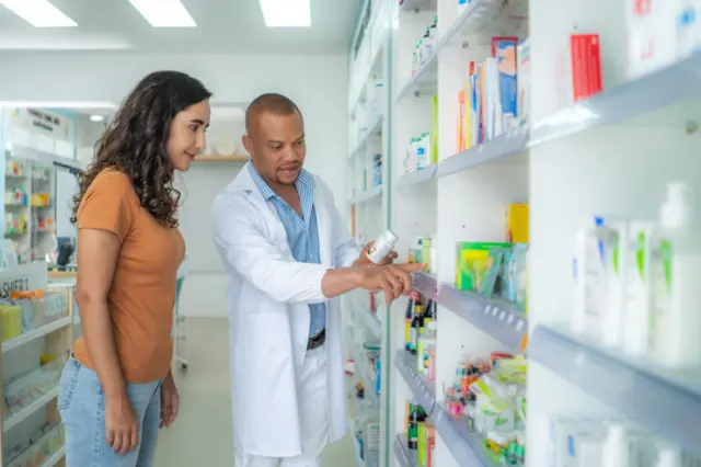 Un farmacéutico con una bata blanca frente a un estante con pastillas y medicamentos recomienda a una mujer que está a su lado un medicamento