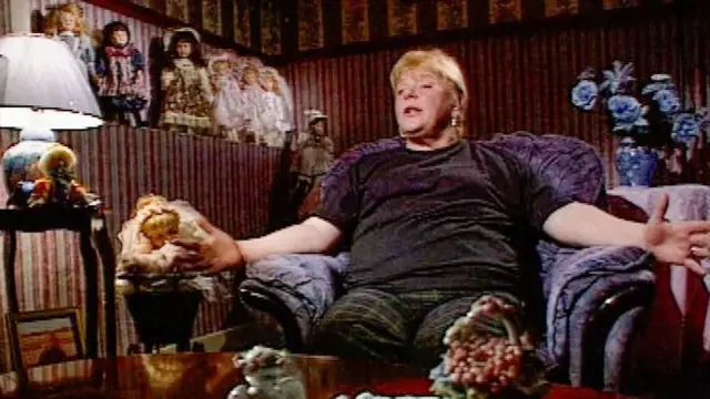 Una imagen televisiva de Haney en su casa, de un documental de principios de la década de 2000. Está sentada en un gran y cómodo sillón morado, rodeada de muñecas infantiles con ropa antigua.