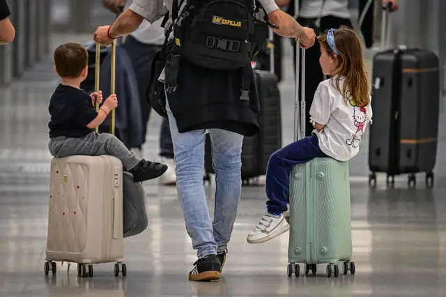Un hombre lleva dos maletas de mano en las que están sentados dos niños.
