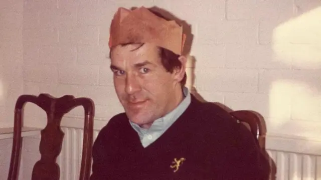 Foto de Ronnie Lockwood, que lleva una típica corona de papel, que se usan en Navidad en Reino Unido. Viste una camisa azul con un suéter de cuello en V color azul marino encima. Tiene ojos azul brillante, cabello castaño y sonríe a la cámara.