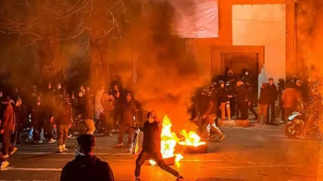 Fuego en la calle de una ciudad iraní y manifestantes con las caras cubiertas.