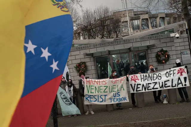 En primer plano, bandera venezolana, de fondo, personas manifestándose con pancartas donde se puede leer en inglés 