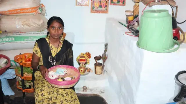 Ankita está sentada en casa sosteniendo una cesta de bambú. Detrás de ella hay bandejas con verduras y las paredes están adornadas con imágenes de dioses.