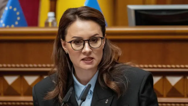 La primera ministra ucraniana, Yulia Svyrydenko, asiste a una sesión del Parlamento ucraniano el 17 de julio de 2025. Viste una chaqueta gris y pantalones cortos azules. Svyrydenko tiene el pelo castaño hasta los hombros y usa gafas marrones. Detrás de ella ondean banderas.