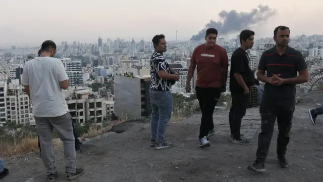Varios hombres observan desde una colina el humo que sale de la emisora estatal iraní tras ser bombardeada por Israel.