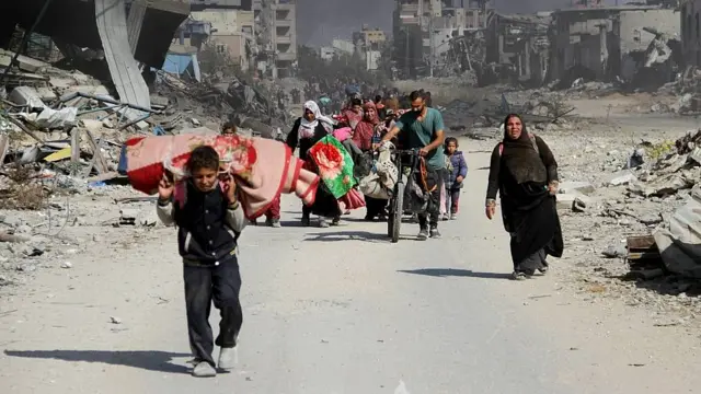 Un niño carga una manta sobre sus hombros junto a familias que se desplazan entre la devastación en Beit Lahia