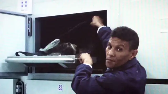 Un asistente de una morgue, vestido de uniforme azul, abrie un cajón refrigerado que contiene un cadáver