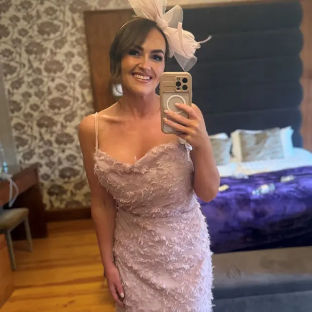 Una mujer en un vestido rosa y un tocado en el pelo posa frente a una cama y se toma un 