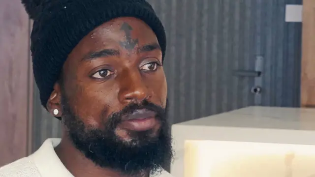 Troy lleva un gorro negro, un cuello color crema, tiene barba, un pendiente y un distintivo tatuaje de ancla en la frente.