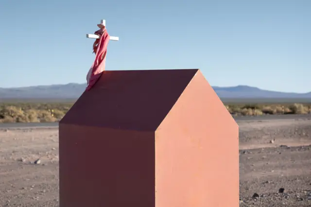 Una caseta de madera con una cruz que tiene una tela envuelta, frente a una carretera desolada