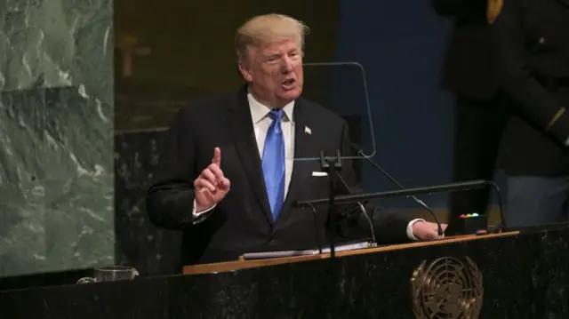 Donald Trump habla ante la Asamblea General de la ONU. Viste un traje oscura, camisa blanca y corbata celeste. Apoya la mano izquierda sobre el podio y tiene la derecha levantada con el dedo índice señalando hacia arriba.